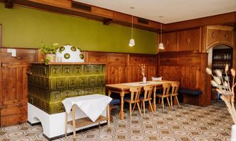 A cozy restaurant with wooden paneling and a tiled stove. The tables are simply set, and the room has a warm, inviting character.