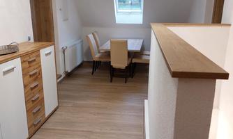 A bright hallway with wooden flooring and modern furniture. On the left, there is a kitchenette, and straight ahead is a dining area with a table and four chairs.