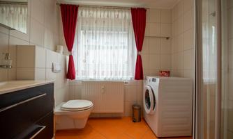 A modern bathroom with orange tiled flooring, a toilet, and a washing machine. Bright windows with white curtains and red drapes create an inviting atmosphere.