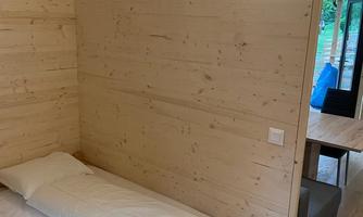 A simple room with a wooden bed and a white bedspread. The walls are made of light wood, and there is a soft light on the wall.