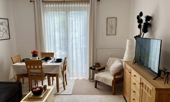 A modern living room with a table, chairs, and a television. The large front window provides plenty of sunlight.