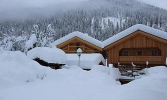 Two wooden chalets surrounded by freshly fallen snow. In the background, there are snow-covered mountains and a forested landscape.