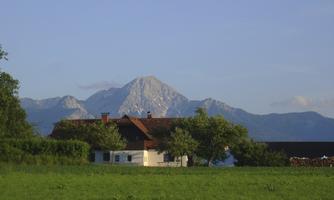 A picturesque scene with a house in the foreground and majestic mountains in the background. The green field contributes to the tranquil atmosphere.