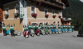 A group of people in green uniforms stands in front of an inn, happily raising their arms. They are gathered on small motorized vehicles.