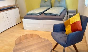 A cozy room with a double bed and blue pillows. There is a round wooden table and an armchair with colorful cushions.