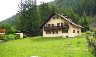 A rural house surrounded by green meadows and trees. In the background, high mountains can be seen.