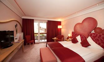 A modern bedroom with red accents and a large, heart-shaped headboard. The room is bright and cozy, with access to a balcony.