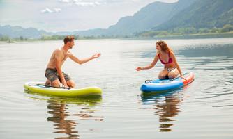 Two people are standing on paddleboards in a calm lake. In the background, the mountains and a clear sky are visible.