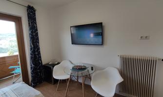 A modern room with a television on the wall. There are two white chairs, a table, and a view of the balcony.
