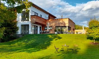 A modern house with wooden siding on a green hill. There is a small table with chairs in the foreground and a beautiful landscape in the background.