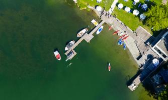 A beautiful lake with several boats at the dock. In the background, there are tables with umbrellas and green meadows.