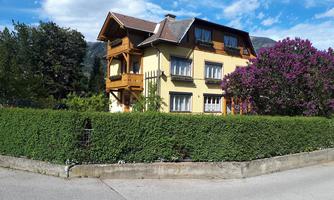 A two-story, yellow house with wooden decorations and a large balcony. Surrounded by a green garden and purple flowering bushes.