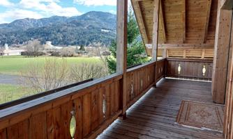 A sunny wooden balcony with a view of green meadows and mountains in the background. The sky is blue with some clouds.
