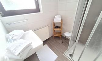 A modern bathroom with a white tiled wall and a shower. Fresh towels are ready on a small shelf.