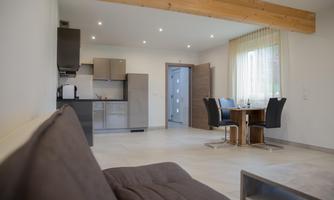 A modern apartment with an open living area and a fully equipped kitchen. The space is brightly lit and features a dining table and chairs.