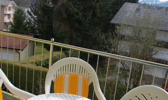 A balcony with two chairs and a round table. In the background, there are trees and a view of the mountains.