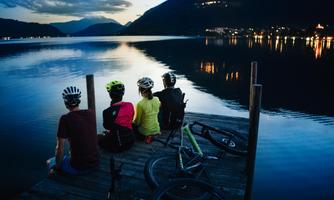 Four cyclists are sitting on a pier by the water, enjoying the evening atmosphere. In the background, mountains and a lit village can be seen.