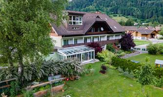 A large house surrounded by trees and a well-maintained garden. In the background, gentle hills can be seen.