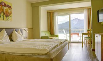 A cozy hotel room with a double bed and a view of the mountains. There is a balcony with a table and chairs, as well as plenty of daylight.
