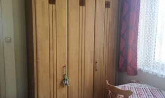 A simple room with a wood-colored wardrobe and a small table. The windows have curtains and the floor is made of wood.