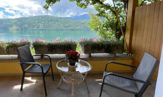 A cozy balcony with two chairs and a table. In the background, a beautiful lake and green mountains can be seen.