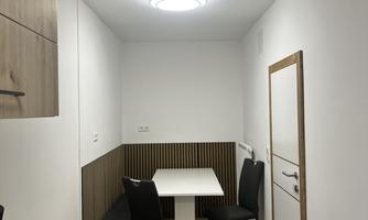 A small, modern room with a table and chairs. On the wall, there are wooden shelves and a bright ceiling lamp.