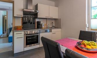 A modern kitchen with a stove, sink, and kitchen utensils. The table is set with a colorful fruit bowl and a piece of tablecloth.