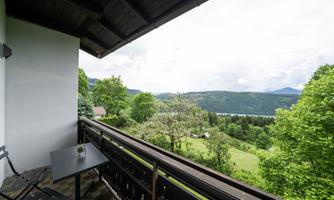 A balcony with a table and chairs offers a wonderful view of green hills and a lake. The sky is cloudy, and the landscape appears calm and inviting.