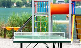 A table tennis table is located in a playground overlooking a lake. In the background, play equipment and green landscapes can be seen.