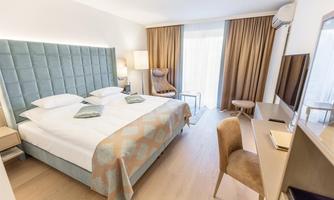 A modern bedroom with a large bed and soft colors. There is a cozy armchair and a desk, as well as plenty of natural light from the windows.
