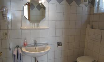 A small bathroom with white tiles and a sink. Next to the sink, there is a toilet and a mirror on the wall.
