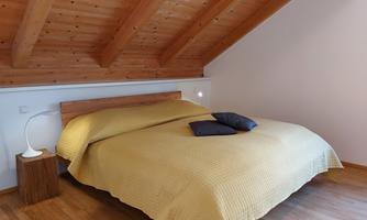 A cozy bedroom with a double bed and wooden ceiling. There is a bedside lamp and two decorative cushions on the duvet.