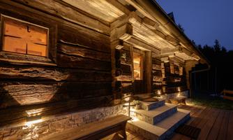 A rustic wooden cabin, illuminated at dusk. The entrance is inviting with stairs and side benches.