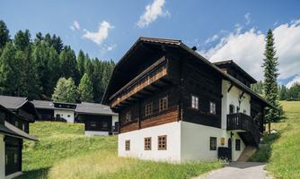 A traditional wooden house in a green, hilly landscape. In the background, more houses and trees are visible.