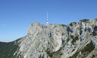 A steep mountain with a rocky surface and a transmission tower at the top. The sky is clear and blue.