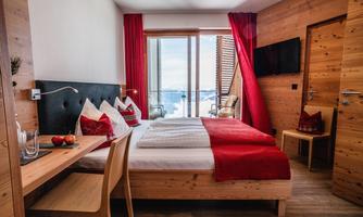 The image shows a cozy hotel room with wooden furniture and red decoration. Large windows offer a view of the outdoor terrace and the landscape.