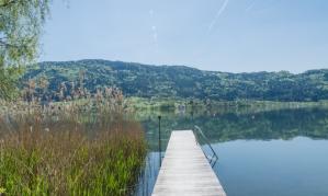 A beautiful pier leads over the calm water of a lake, surrounded by green banks and gentle hills. The sun shines bright in the clear blue sky.