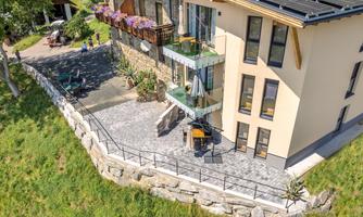 A modern house with several floors and a balcony. The garden is beautifully landscaped and there is a terrace with stones.