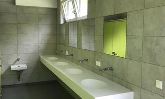 A modern bathroom with gray tiles and a long sink. There are large windows and a green door.