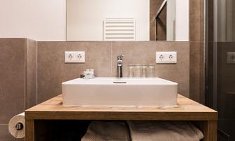 A modern bathroom with a wooden washbasin and a white wash bowl. Towels are neatly stacked and a glass door leads to the shower.