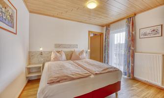 A cozy bedroom with a large double bed and warm wooden interiors. There is a window with curtains and access to the balcony.
