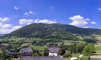 A picturesque landscape with green hills and a clear blue sky. In the foreground, houses can be seen, surrounded by nature.