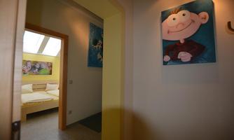 A bright hallway with two pictures on the wall and a view of a bedroom. The bedroom has a large window and bright bedding.