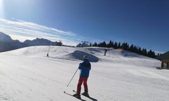 A skier is standing on a snow-covered slope under a clear blue sky. In the background, the mountains and a ski lift are visible.