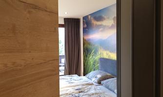 Insight into a modern bedroom with a large bed and a landscape wallpaper. The room features light colors and windows with curtains.