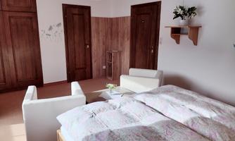 A simple room with a double bed, a white sofa, and a small table. The walls are light, and there are wooden cabinets.