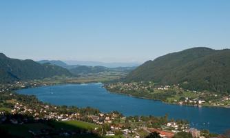 A picturesque landscape with a tranquil lake, surrounded by green mountains. In the foreground, small houses and meadows are visible.