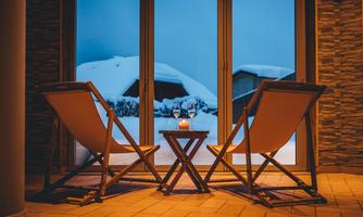 Two lounge chairs are in front of a large window overlooking a snow-covered landscape. A candle holder and two glasses are on a small table between the chairs.