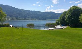 A calm lake surrounded by mountains and green grass. Boats are moored at the shore and the sky is partly sunny.