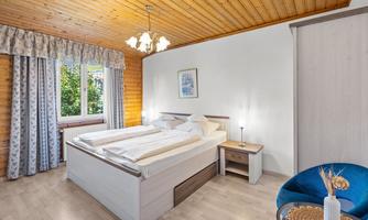 A cozy bedroom with a large bed and wooden ceiling. Daylight comes through the window and there is a modern seating area.